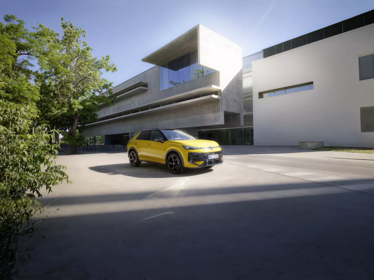 Ryškiai geltonas naujasis Volkswagen T-Roc modelis važiuoja saulėta, modernia, architektūrine miesto aplinka.