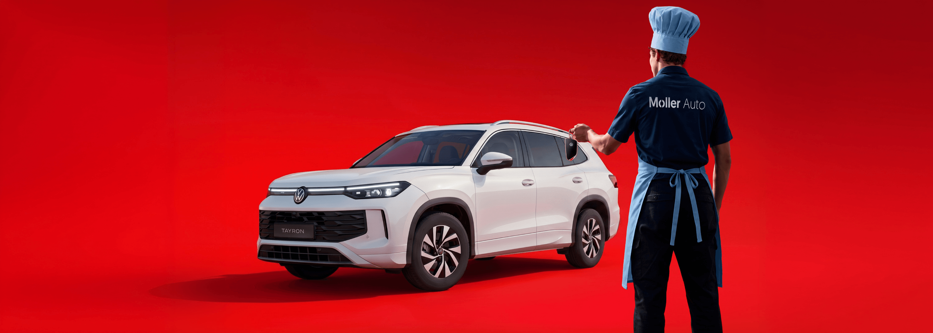 Baltas Volkswagen Tayron visureigis raudoname fone, šalia Möller Auto darbuotojas su virėjo kepure ir prijuoste, rankoje automobilio raktelis.