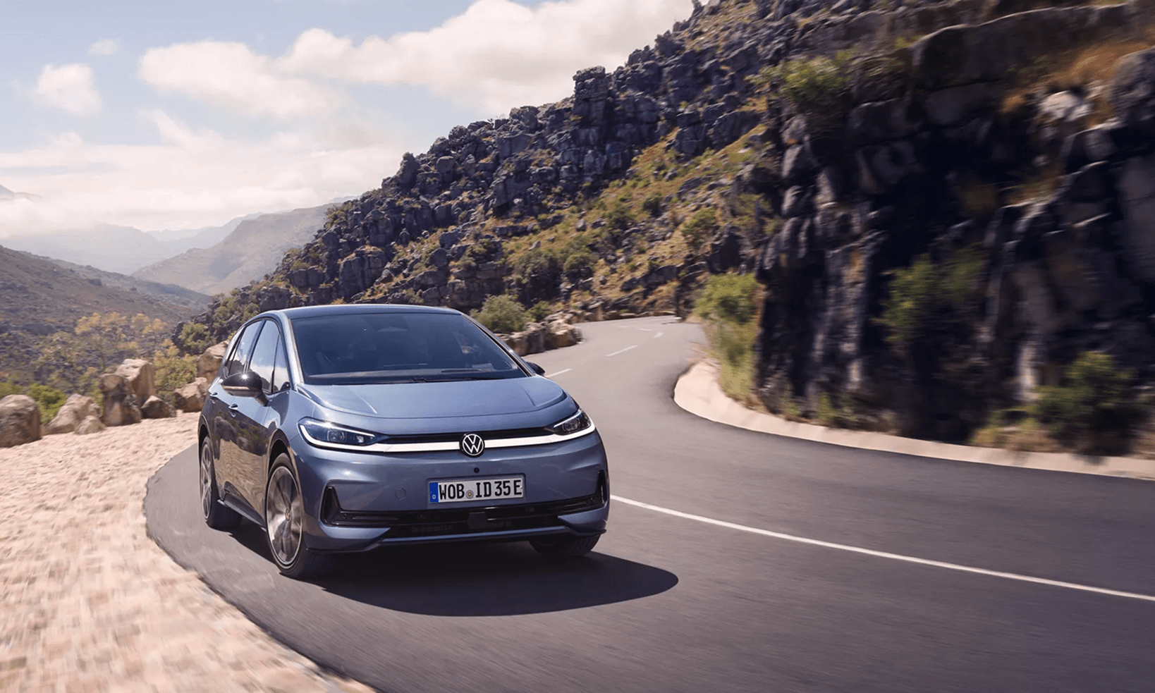 Mėlynas Volkswagen ID.3 NEO elektromobilis pastatytas kalnų apžvalgos aikštelėje po medžiu, fone du žmonės grožisi kalnų peizažu.